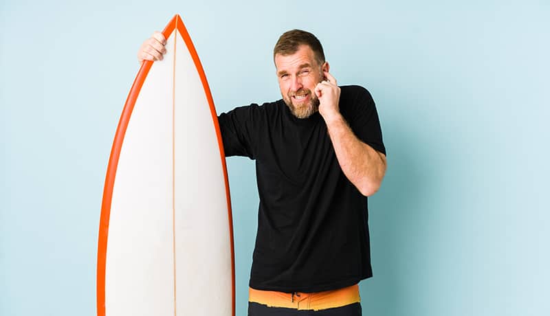 Mann mit Surfbrett fasst sich ans Ohr, weil er an einer Gehörgangsexostose leidet.